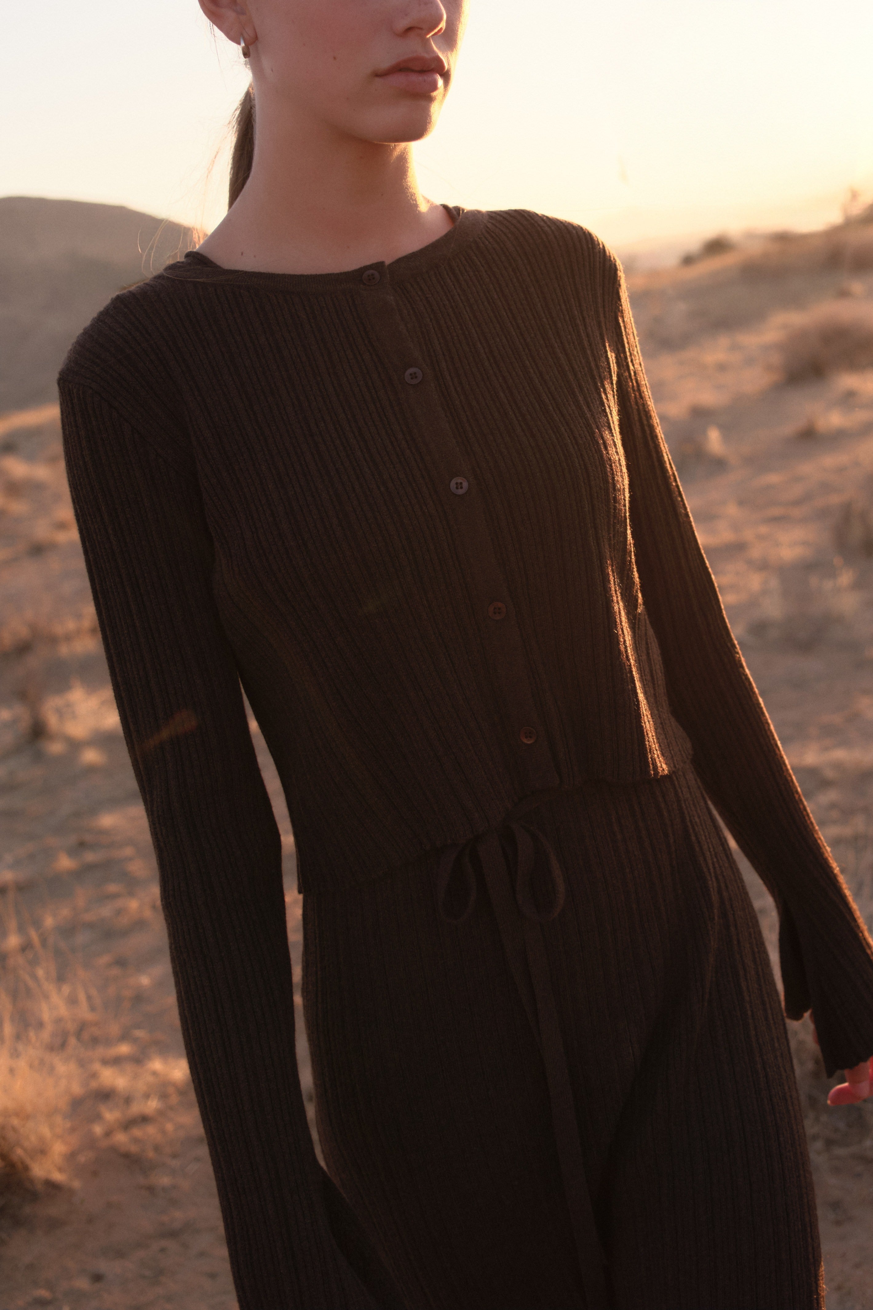 Close-up of a model wearing a black ribbed button-up top with matching drawstring pants, standing in a desert landscape at sunset.