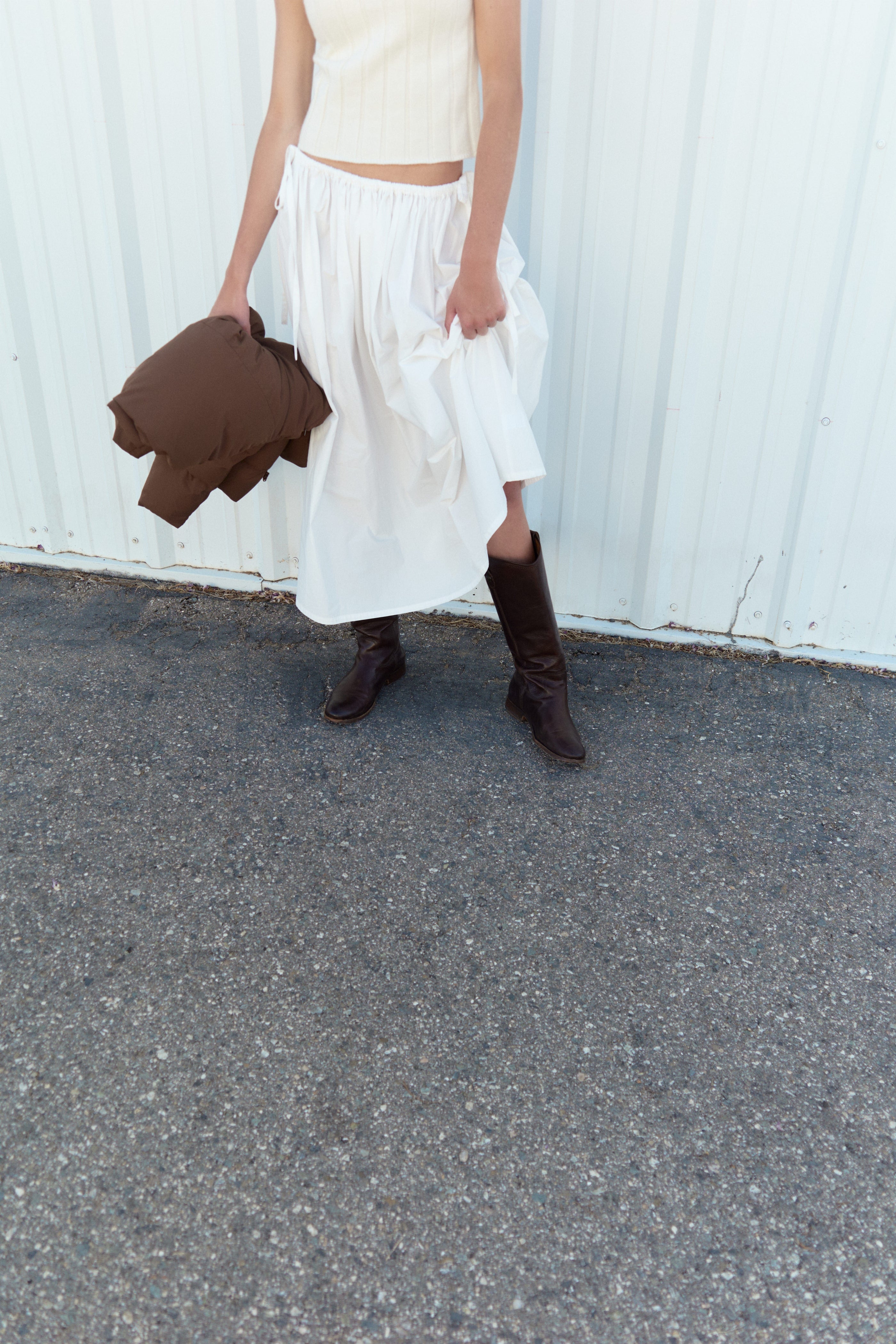 A person wearing a white sleeveless top, flowy white skirt, and tall brown boots stands on asphalt, holding a brown jacket, with a white metal wall in the background. Their face is not visible.
