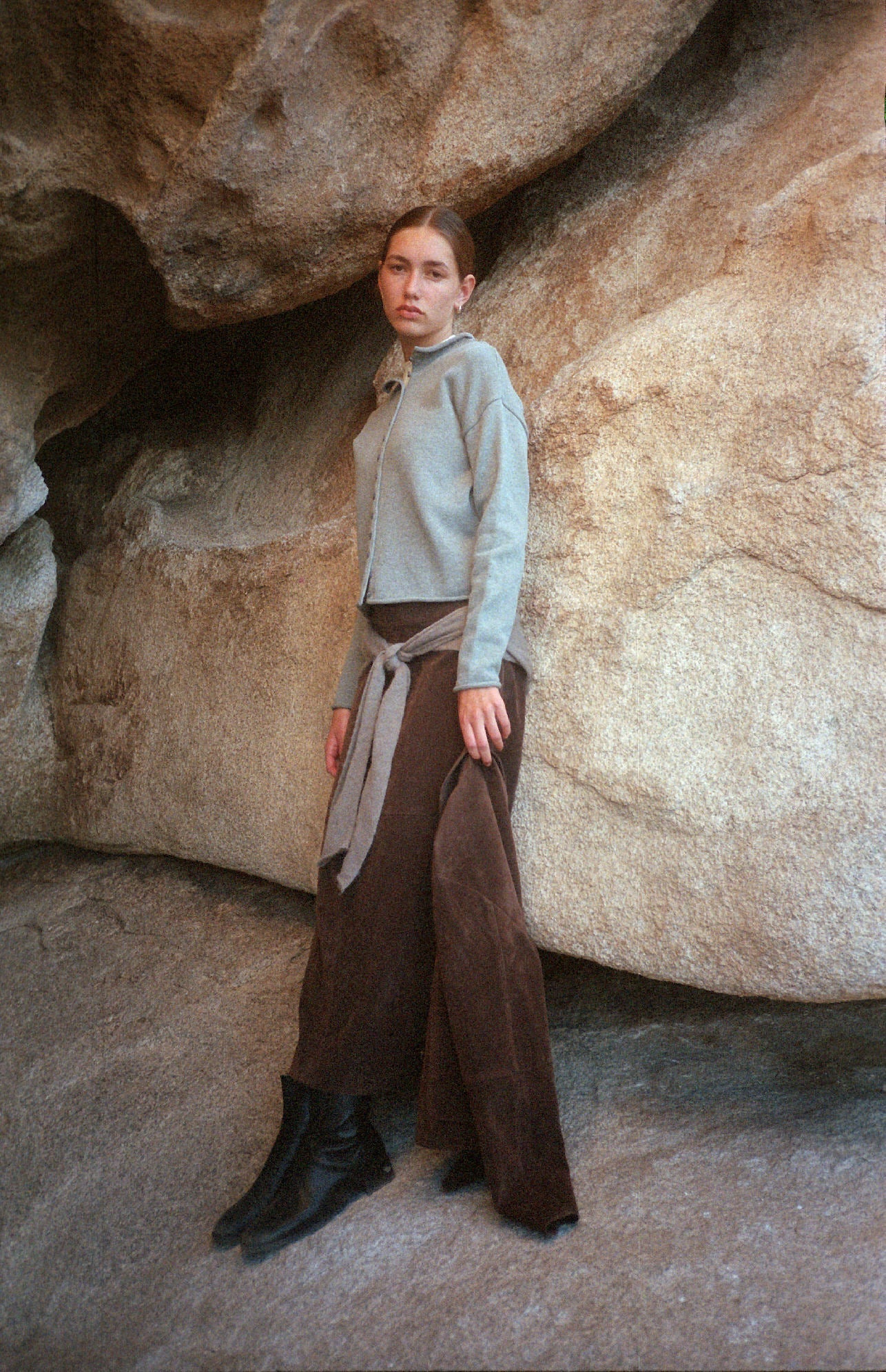 A woman with straight hair stands against large beige rocks, wearing a light gray zip-up jacket, a gray sweater tied at her waist, brown wide-leg pants, and black boots, looking calmly at the camera.