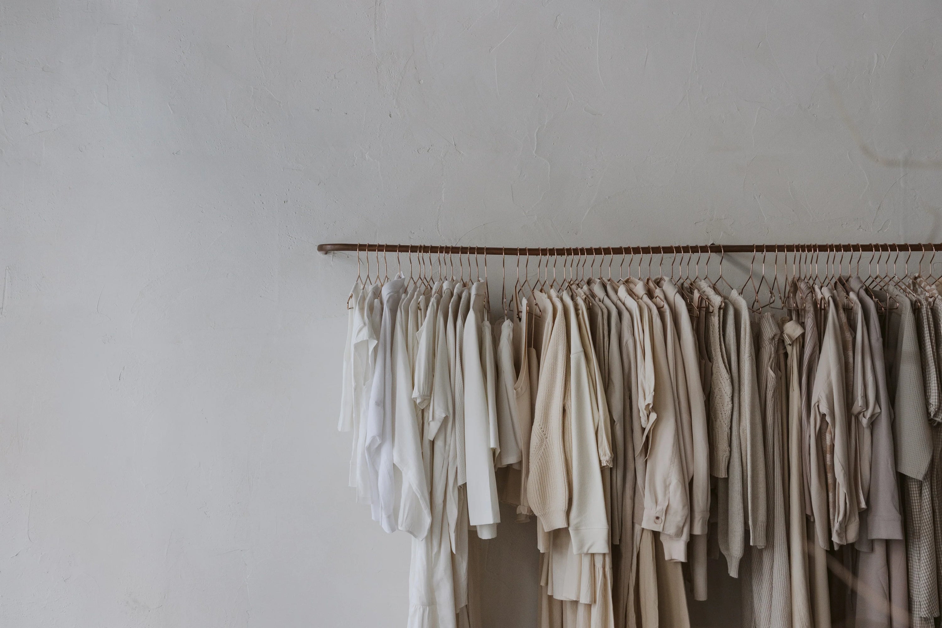Minimalist clothing rack against a textured white wall, featuring a curated assortment of garments in soft neutrals—ivory, beige, taupe, and sand. The tonal palette and natural fabrics reflect a refined, timeless aesthetic.