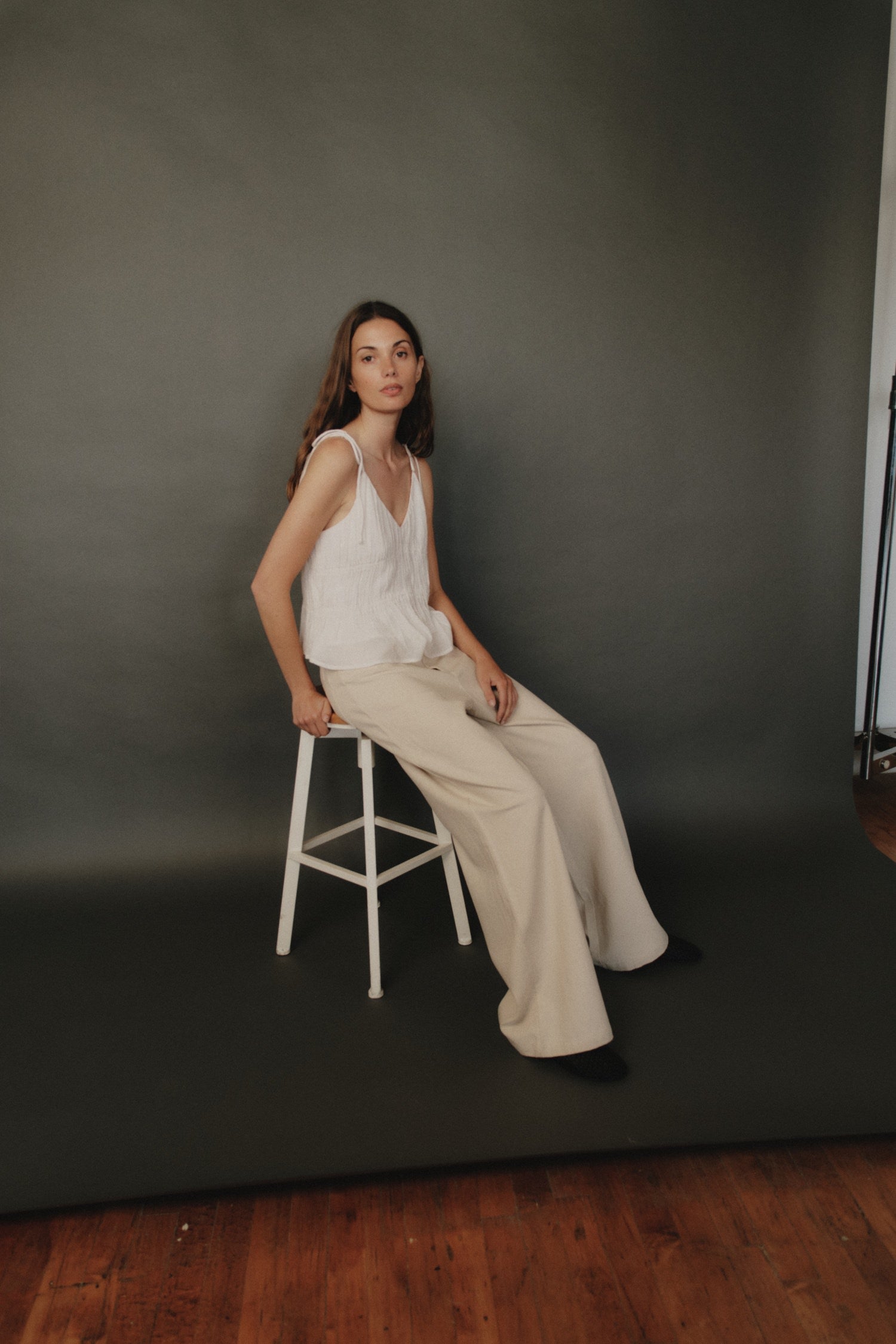 Woman seated on a white stool against a dark backdrop, wearing a sleeveless white top and wide-leg beige trousers.