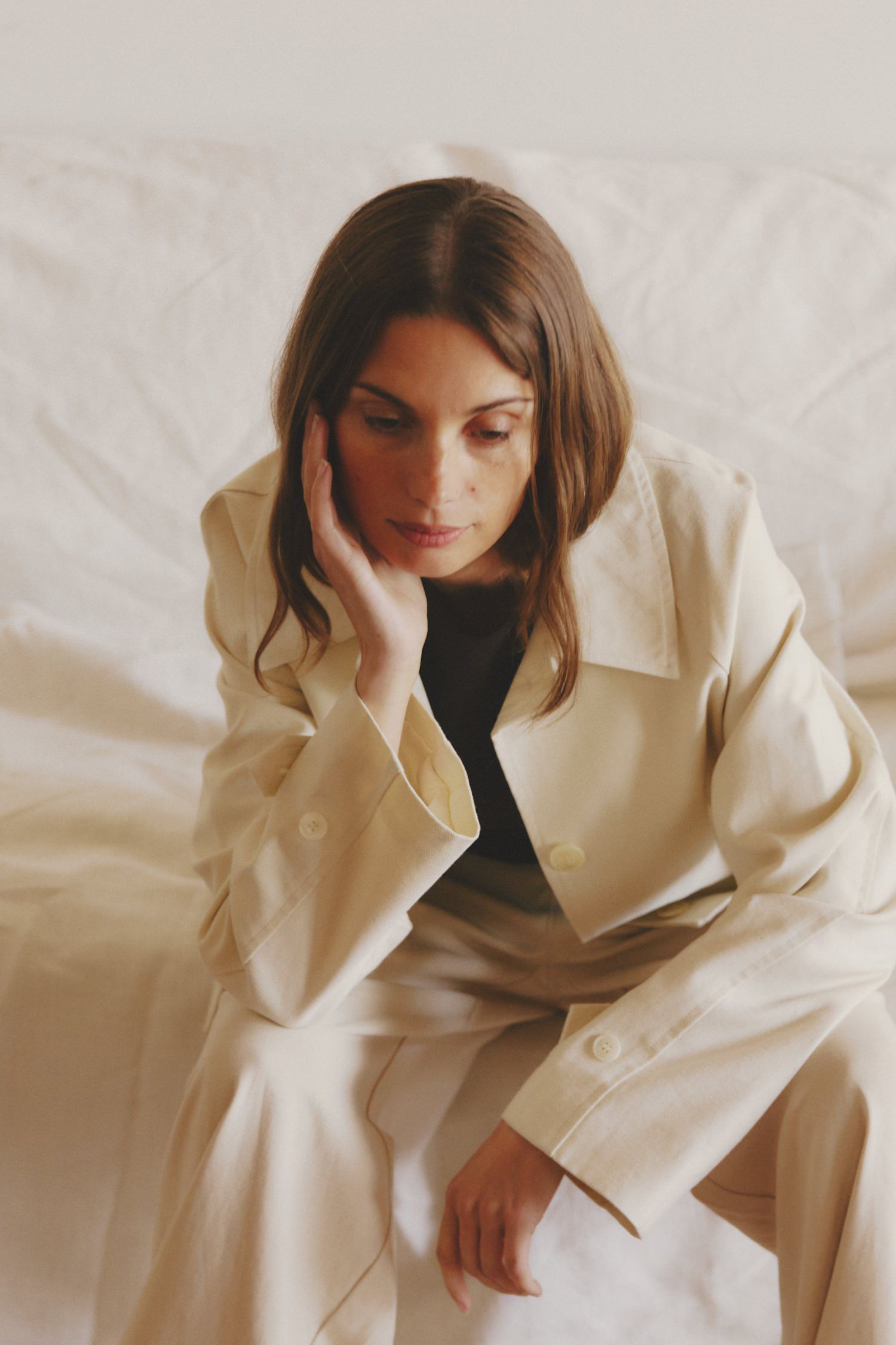 Woman in a cream-colored suit sitting on a white couch, resting her face on her hand in a thoughtful pose.