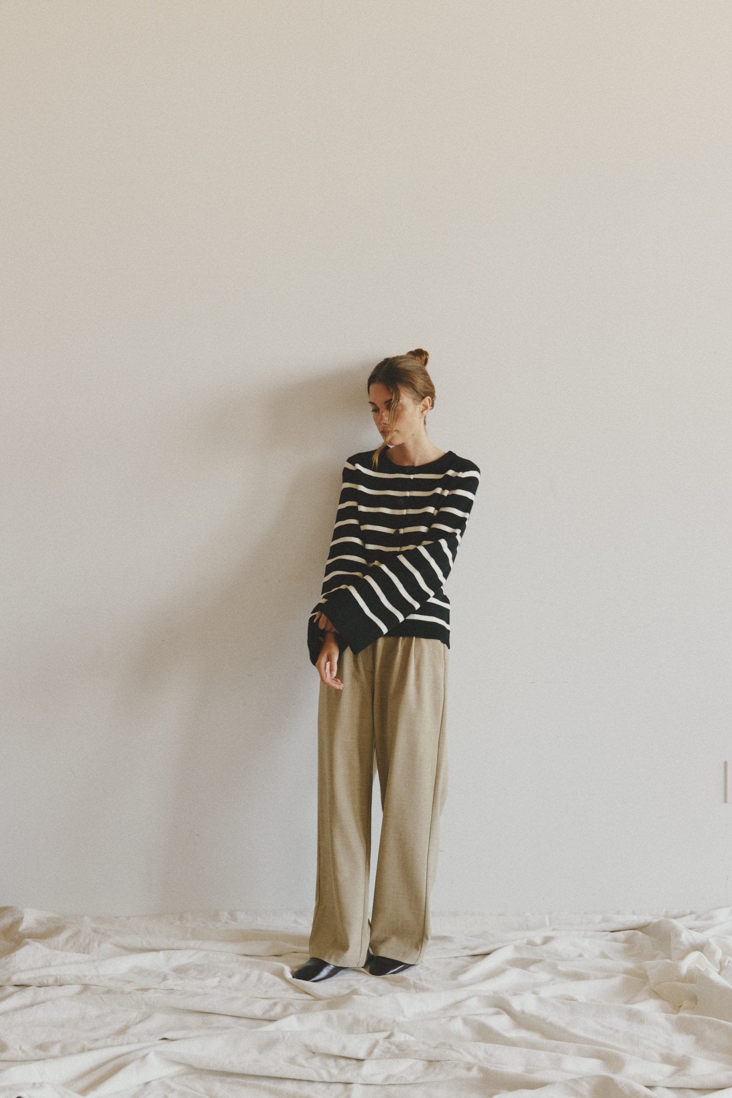 Woman in a black and white striped sweater and beige trousers standing against a plain wall on a draped white sheet.