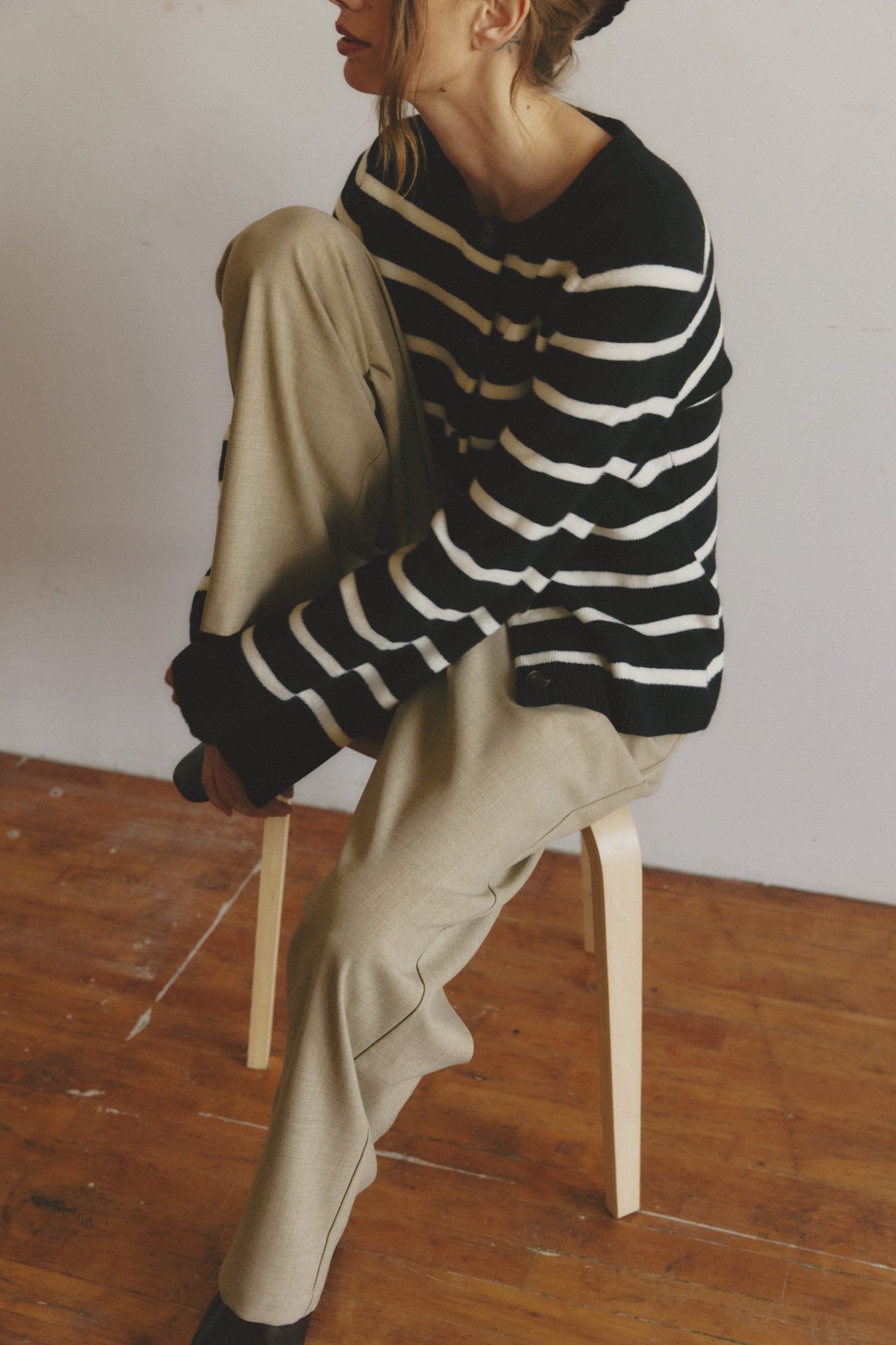 Woman seated on a stool wearing a black and white striped sweater and beige pants, resting one knee up.
