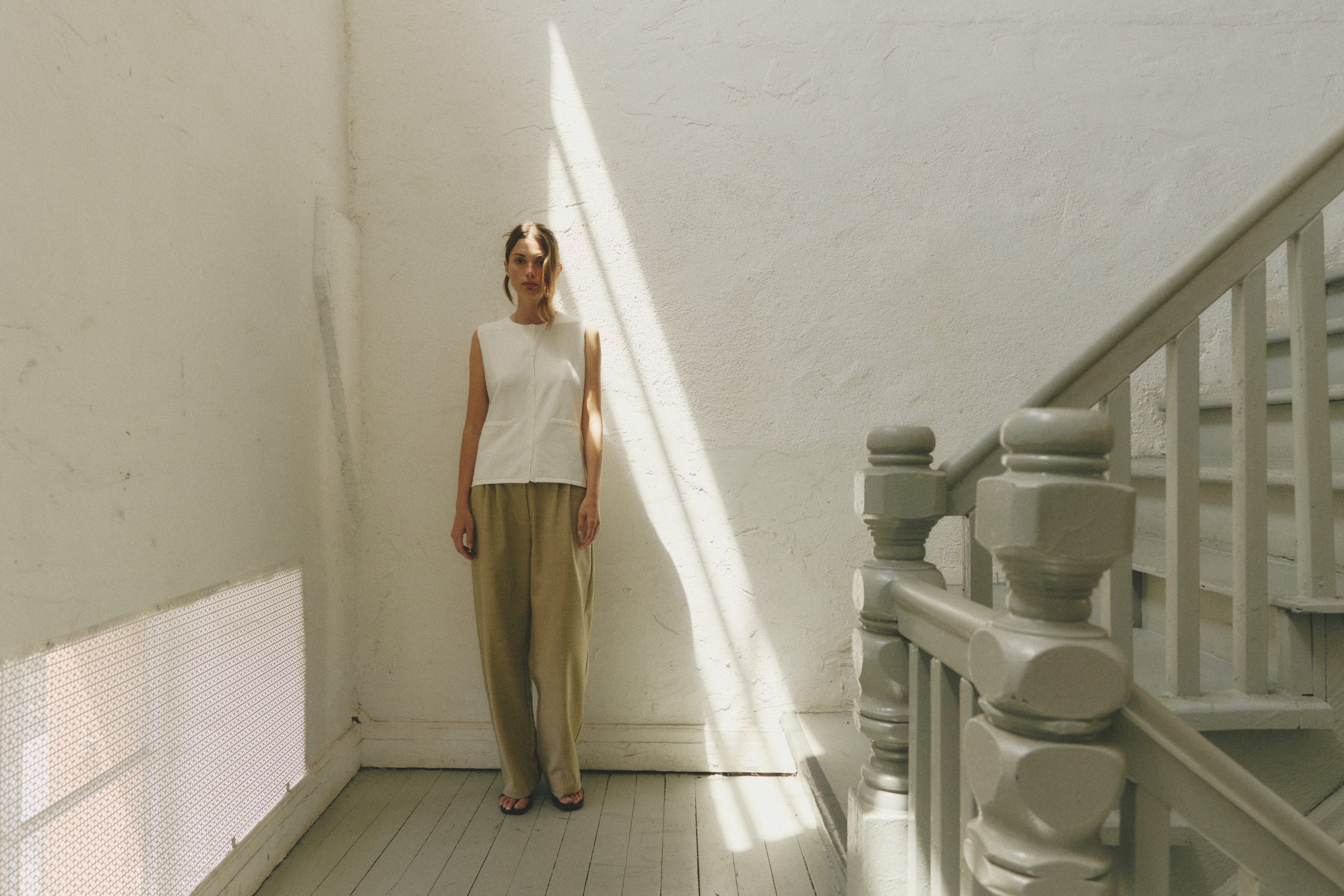 Woman standing in a sunlit stairwell, wearing a sleeveless white top and beige pants.
