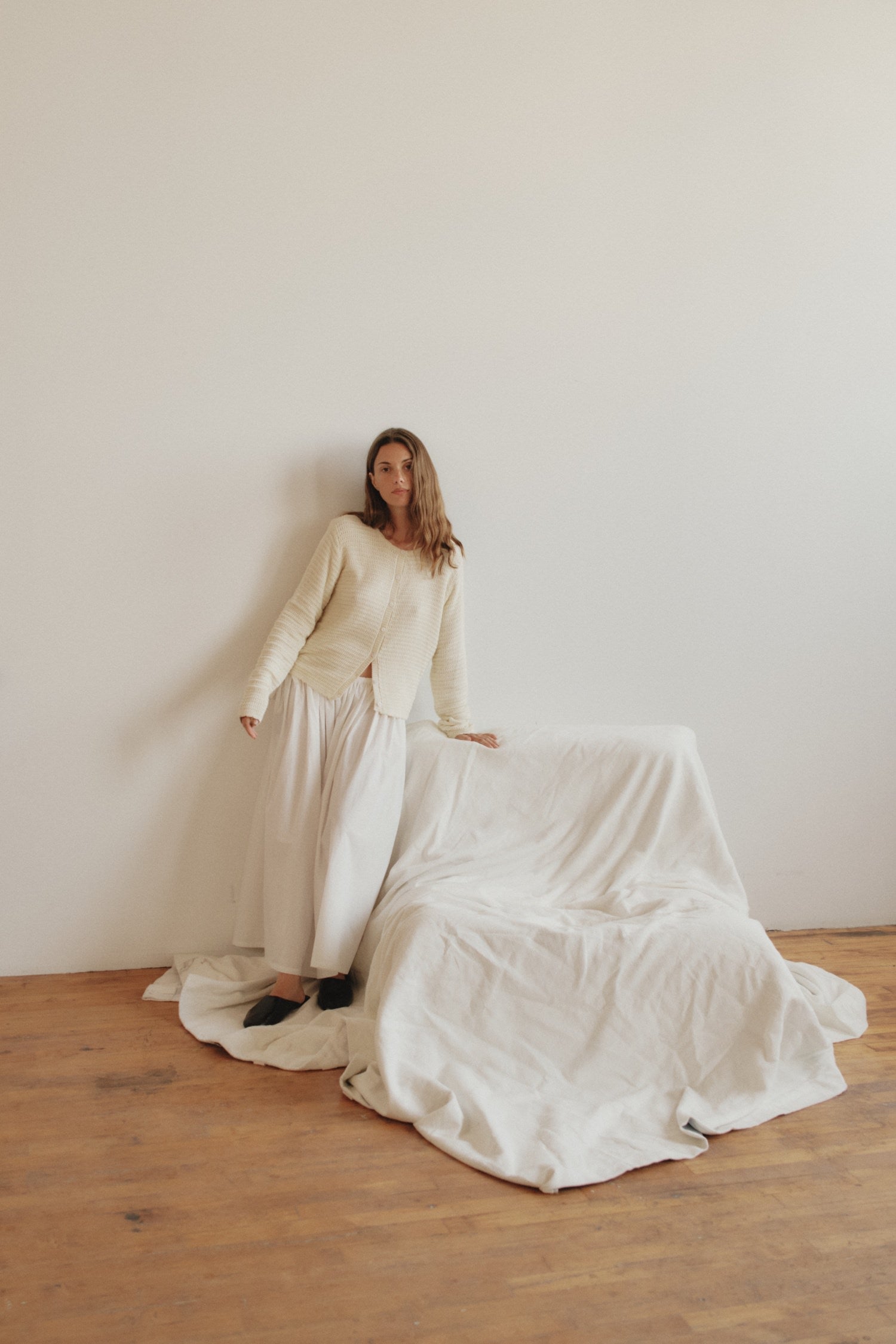 Woman standing beside a white sheet-draped couch, wearing a light sweater and flowing skirt in a minimal, sunlit room.