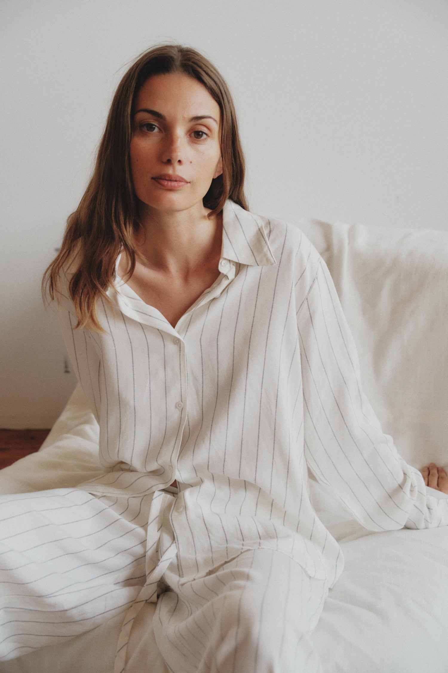 Woman seated on a white couch, wearing a relaxed white outfit with thin vertical stripes, looking calmly at the camera.