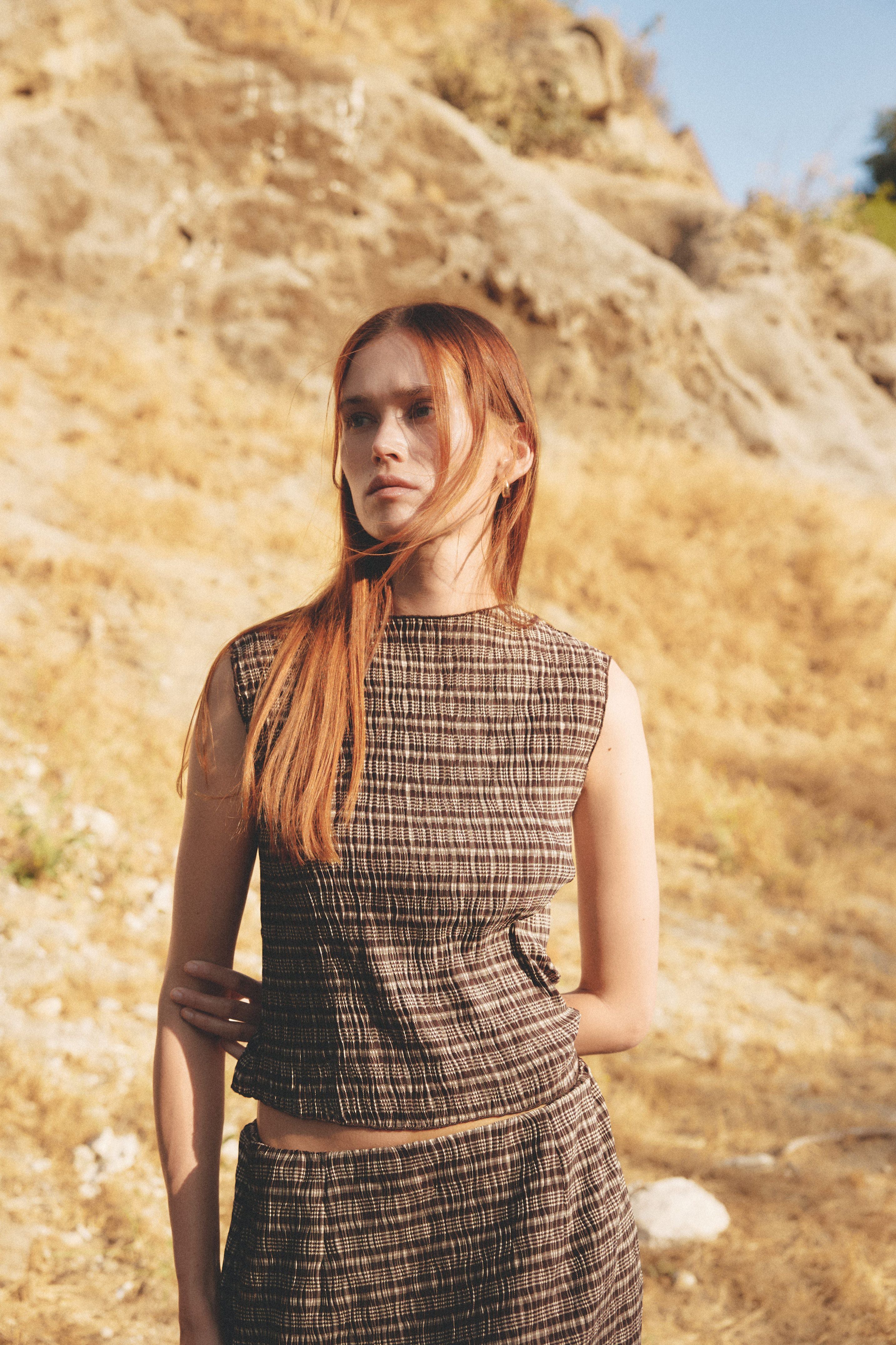 Woman standing against a sunlit hillside in a brown plaid sleeveless top and matching skirt. The warm, dry landscape and natural lighting highlight the earthy tones and textured fabric, evoking a grounded, late-summer feel.