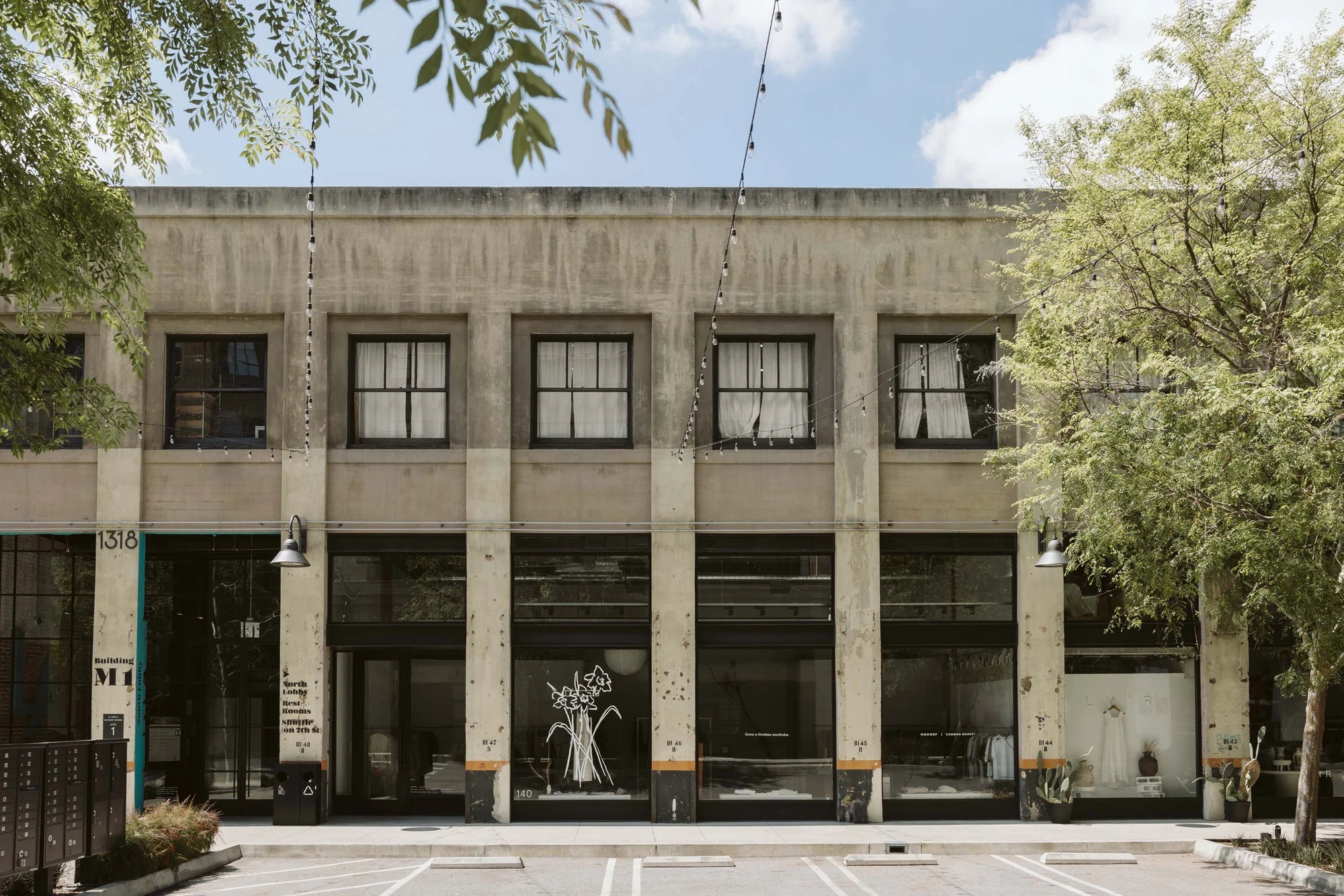 Street view of a modern storefront with tall concrete columns and large industrial windows. The space features minimal signage, with delicate floral illustrations on the glass and visible clothing racks inside, reflecting Mod Ref’s refined, architectural retail presence.