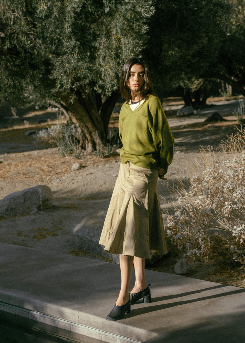 A woman with straight hair stands against large beige rocks, wearing a light gray zip-up jacket, a gray sweater tied at her waist, brown wide-leg pants, and black boots, looking calmly at the camera.