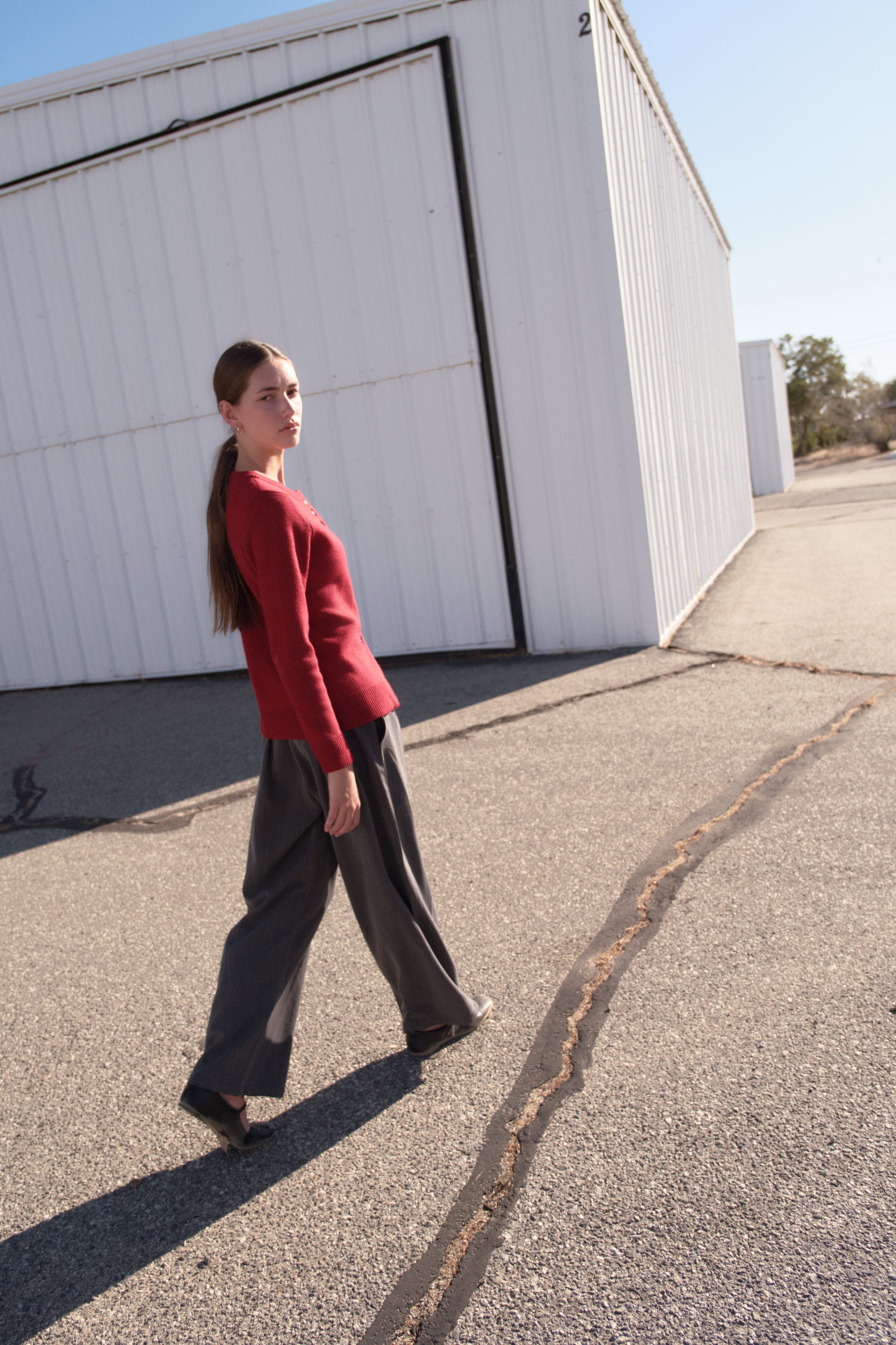 Model walking outdoors near white industrial buildings, wearing a red long-sleeve top and wide-leg charcoal pants, styled for a minimalist fall look.