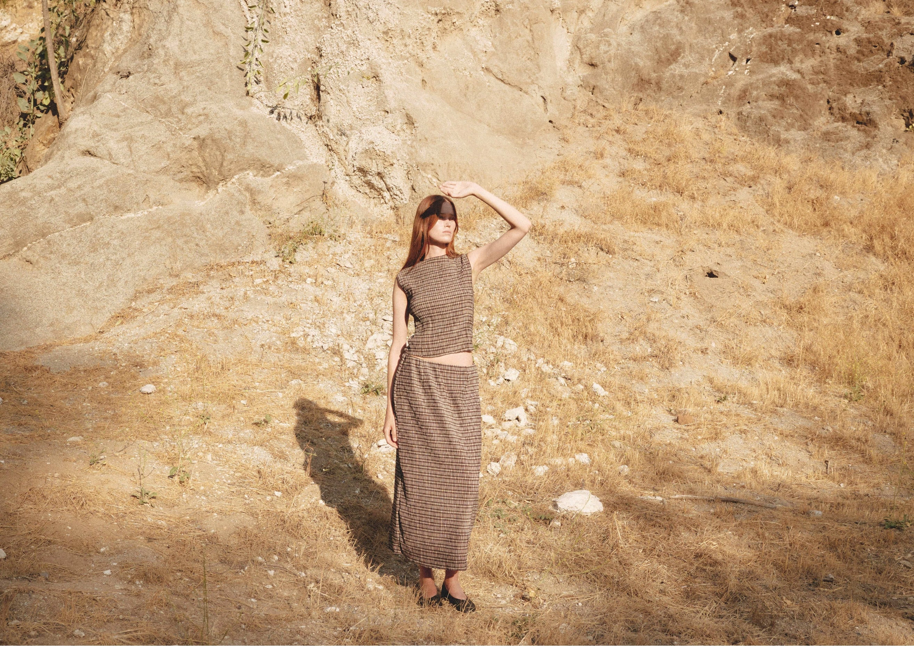 Woman standing on a dry hillside in a brown plaid two-piece set, hair loose, with golden light casting shadows on the ground.