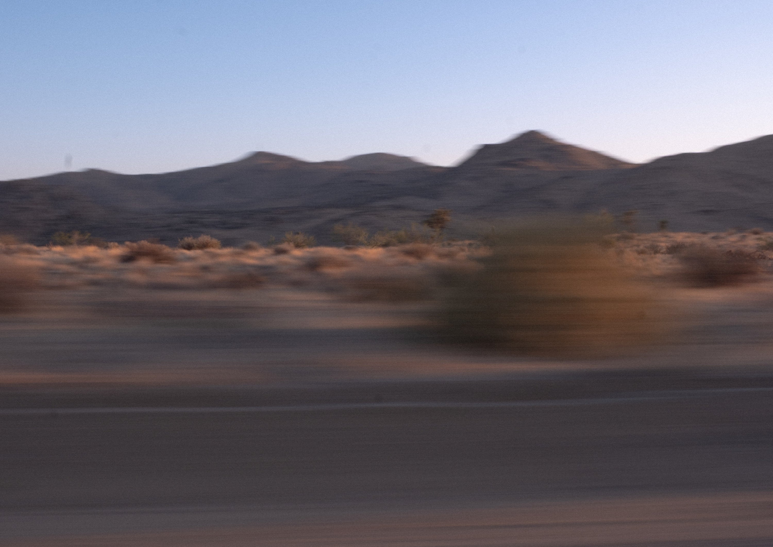 Blurred desert landscape captured in motion, with dry brush and mountains in the background under a clear evening sky.