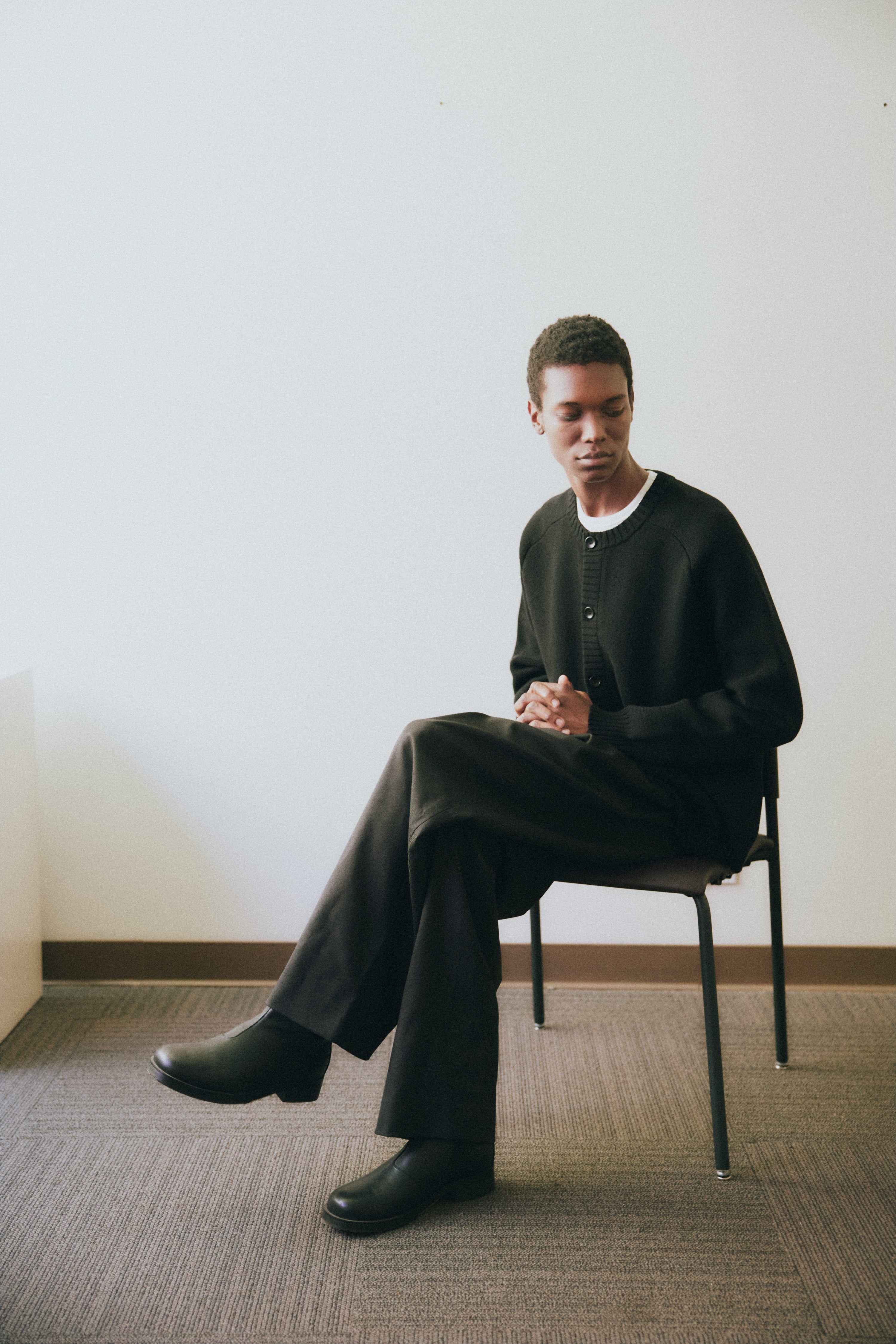 Man seated on a simple chair in a minimal studio space, wearing a black buttoned cardigan, wide-leg trousers, and black boots. His posture is composed, and the soft lighting emphasizes the clean lines and relaxed tailoring.