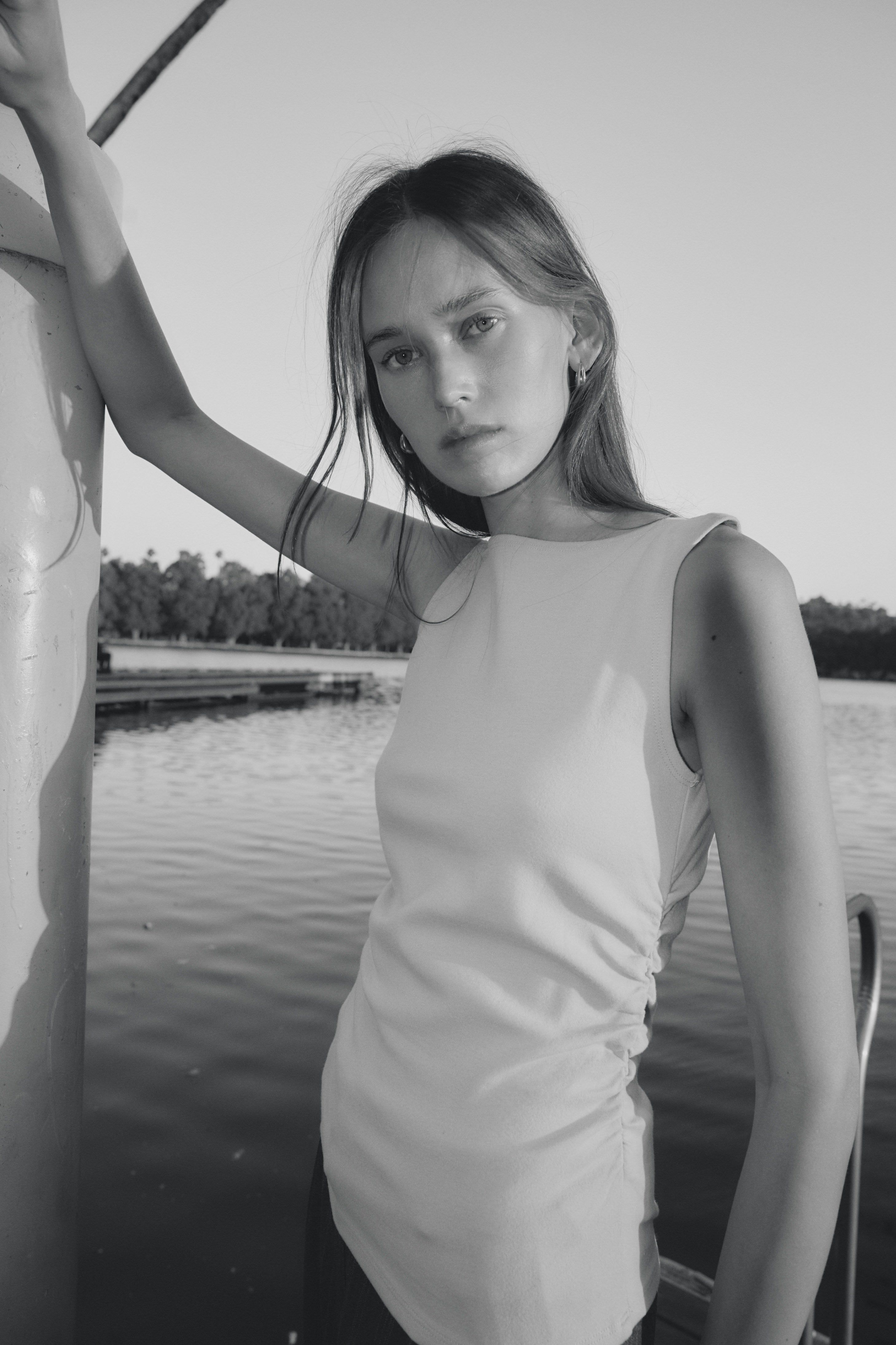 Woman in sleeveless top leaning on a pole by a lakeside, gazing at the camera in black and white photo.