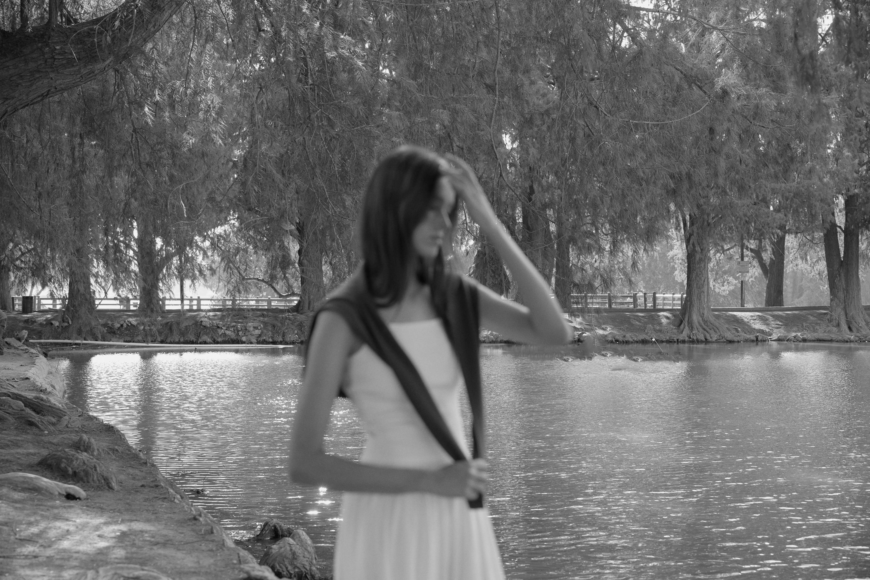 Blurred woman standing near a lakeside path, surrounded by tall trees in a peaceful black and white forest scene.