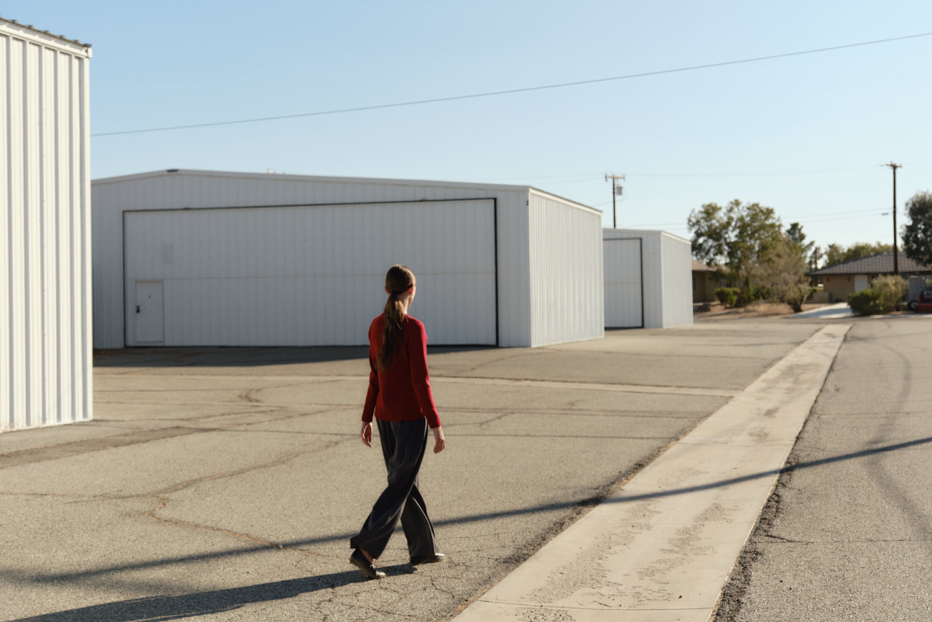 Model walking through an industrial outdoor space with white metal buildings, wearing a red long-sleeve top and dark wide-leg pants, casting a long shadow on the pavement.