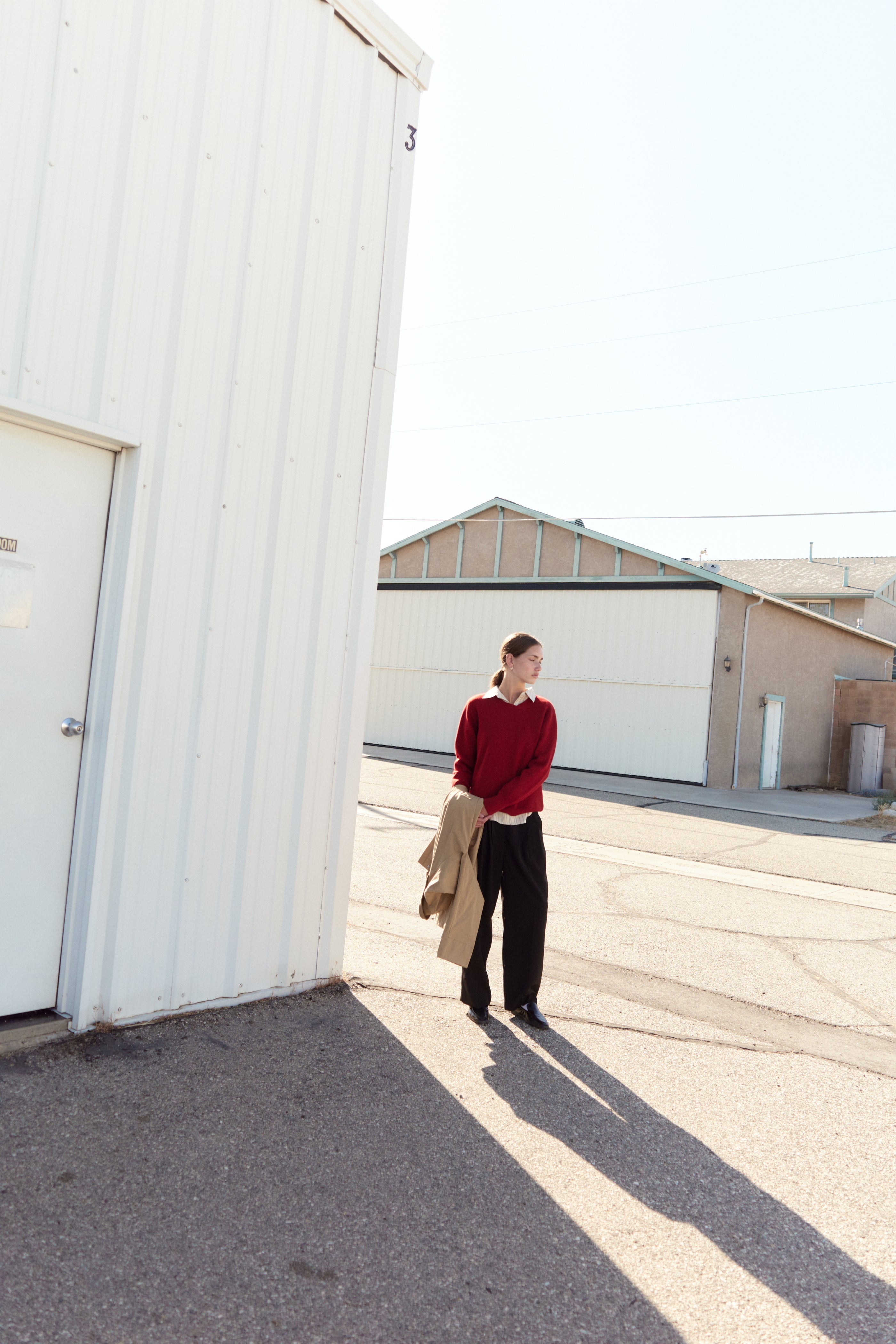 Model standing outside near white industrial buildings, wearing a red sweater over a collared shirt and black wide-leg trousers, holding a tan coat in her hand with long shadows cast on the pavement.