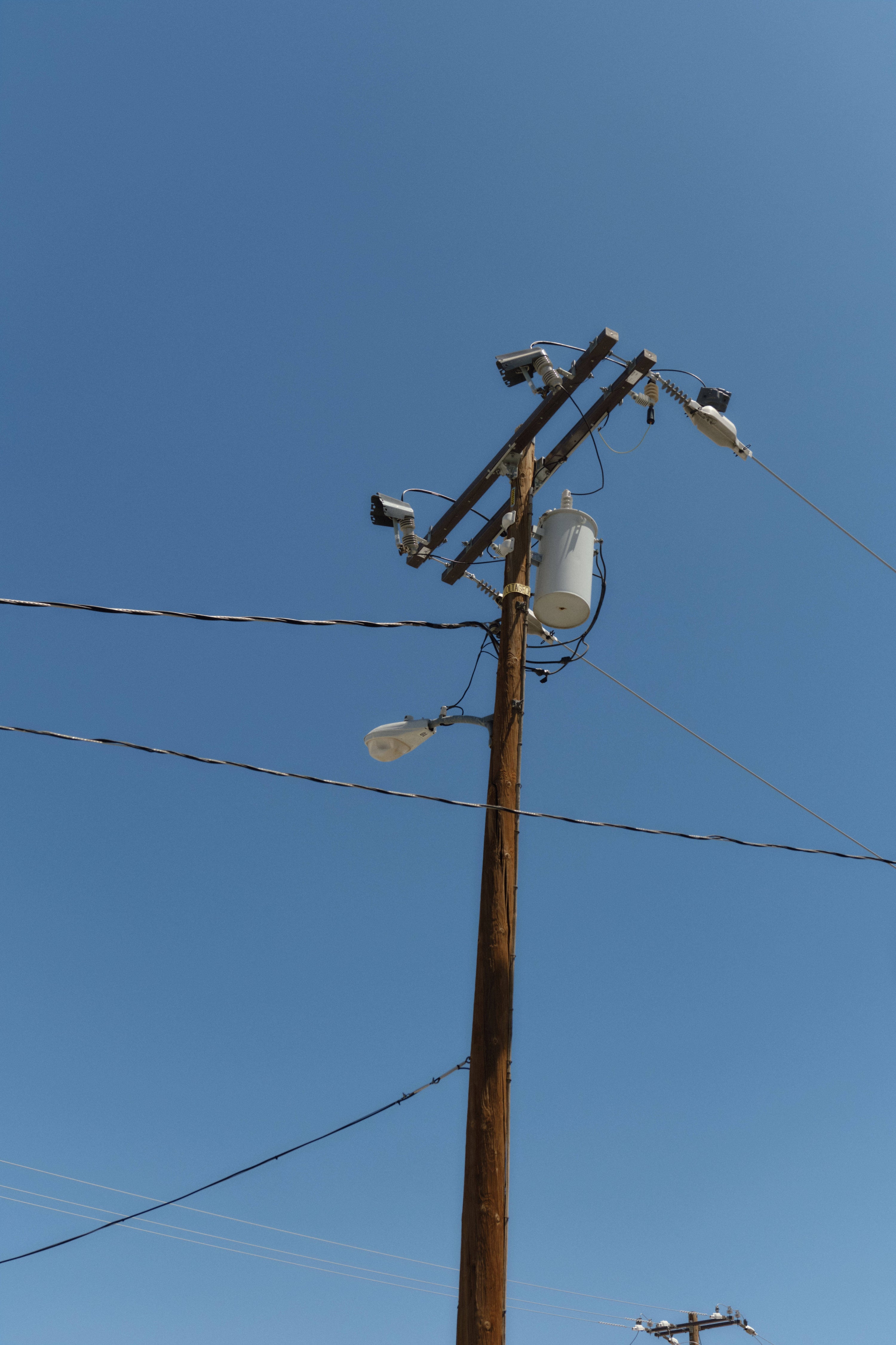 Wooden utility pole with power lines, transformers, and security cameras against a clear blue sky.