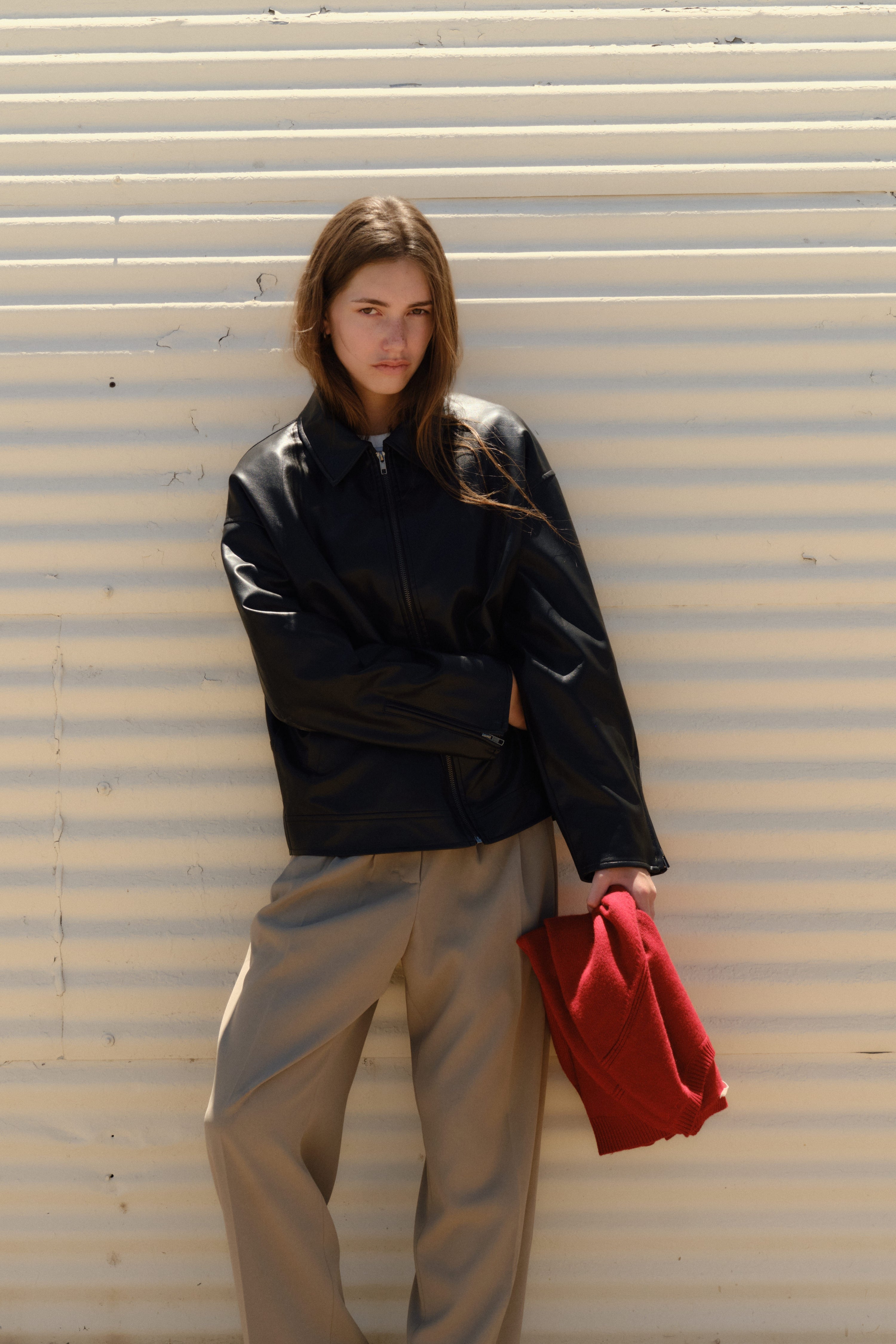 Person wearing a black jacket and beige pants holding a red bag against a corrugated metal background