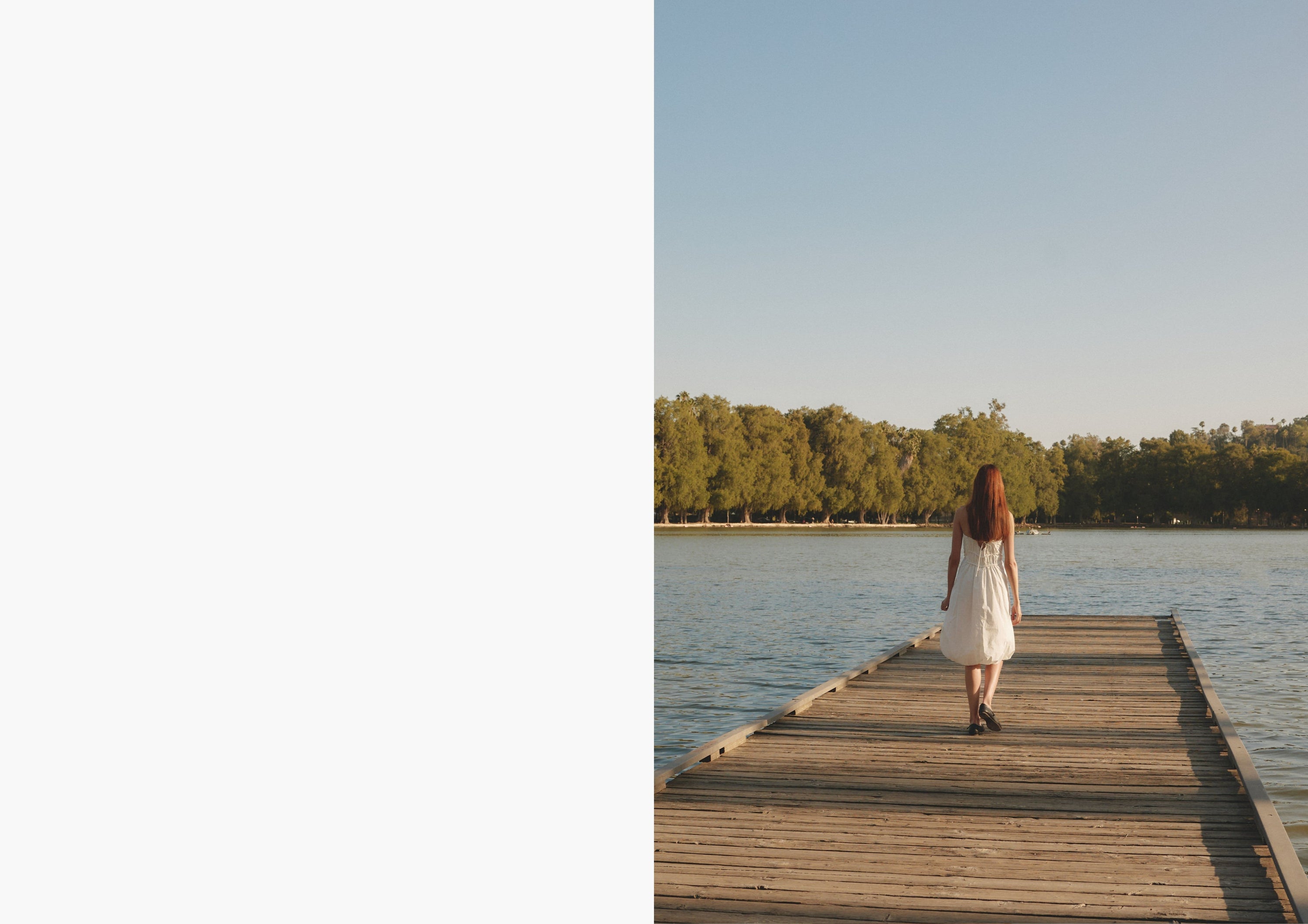 Back view of a woman in a white dress walking alone down a wooden dock toward the lake, framed by a peaceful, wooded backdrop.