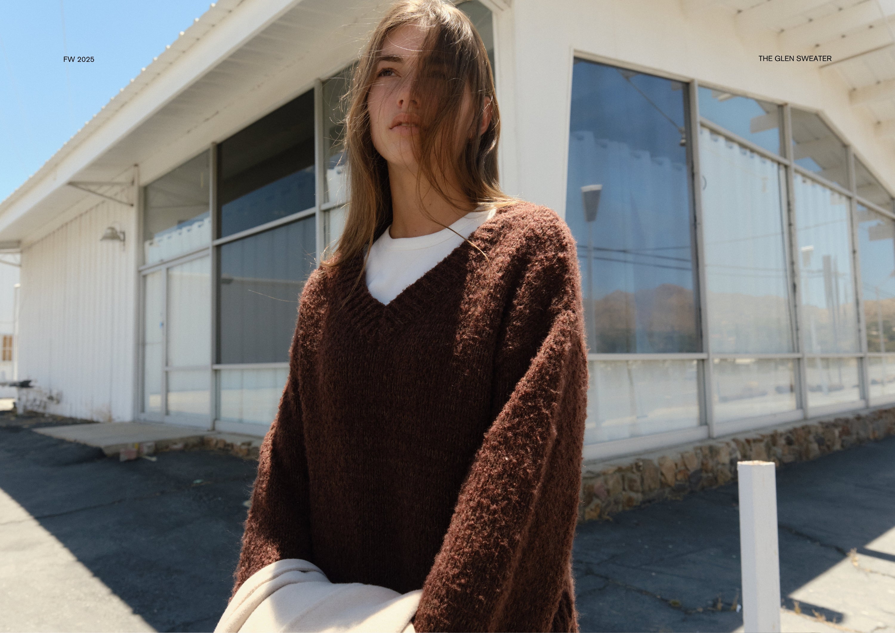 Close-up of a model standing outside a white building, wearing a textured dark brown V-neck sweater layered over a white top, holding a light coat in her arms with hair blowing gently in the wind.