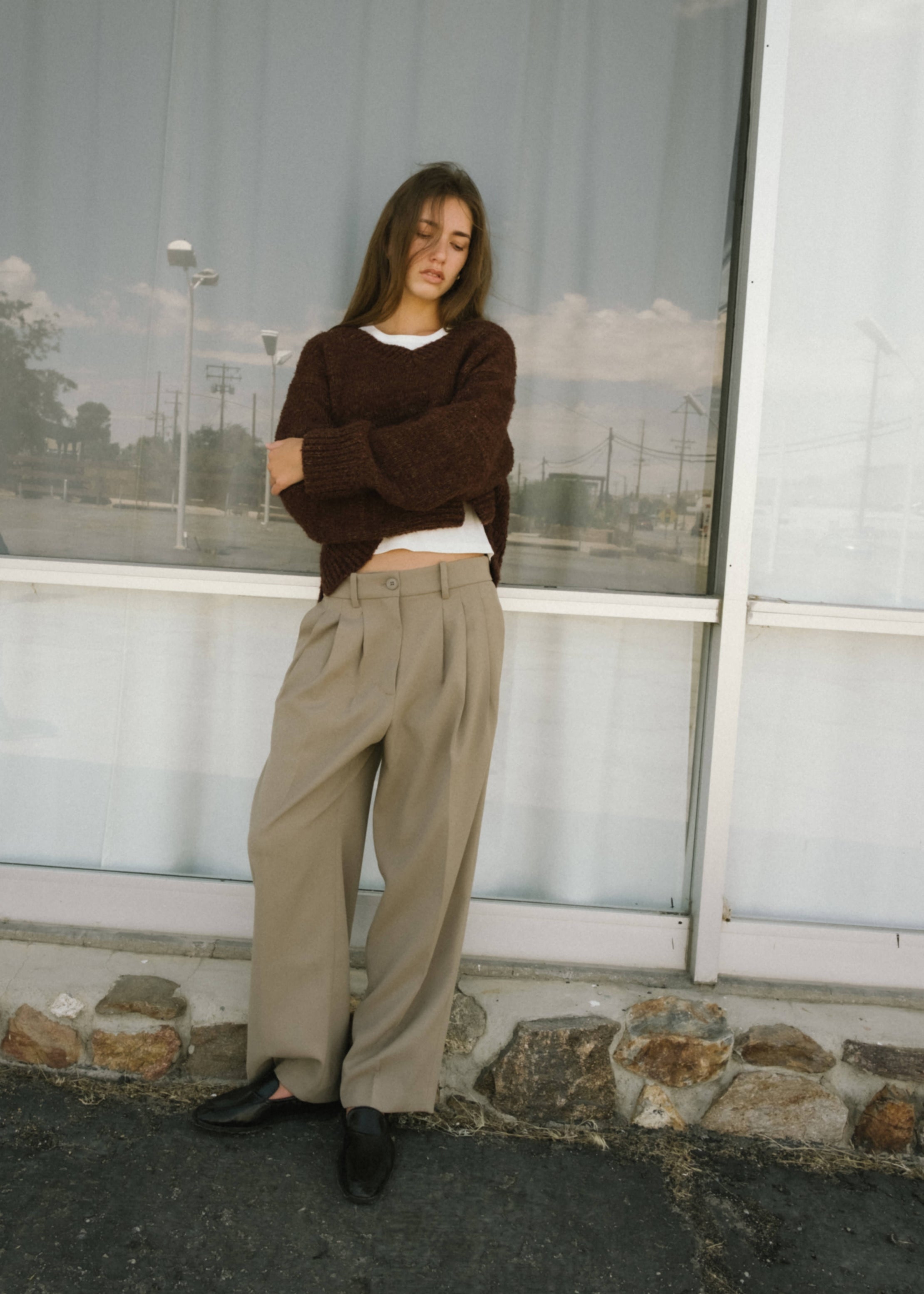 Model leaning casually against a large window, wearing a dark brown cropped sweater layered over a white top, paired with high-waisted, loose beige trousers and black shoes.