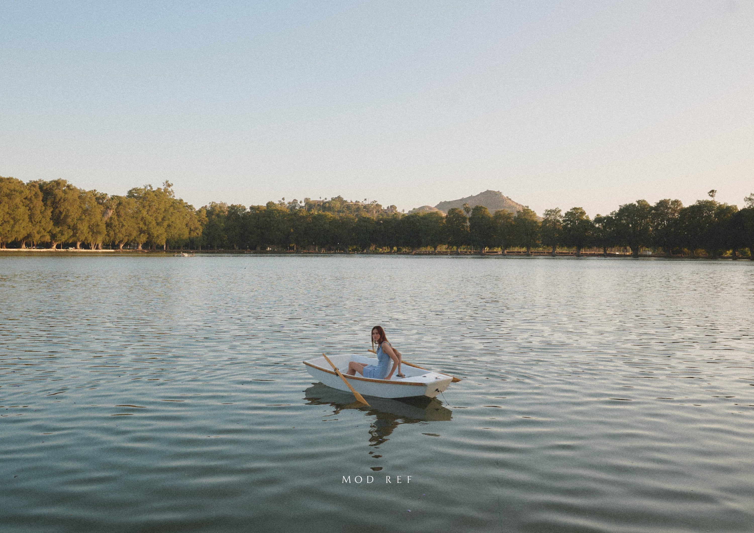 Woman in a rowboat on a calm lake with trees and hills in the background during sunset.