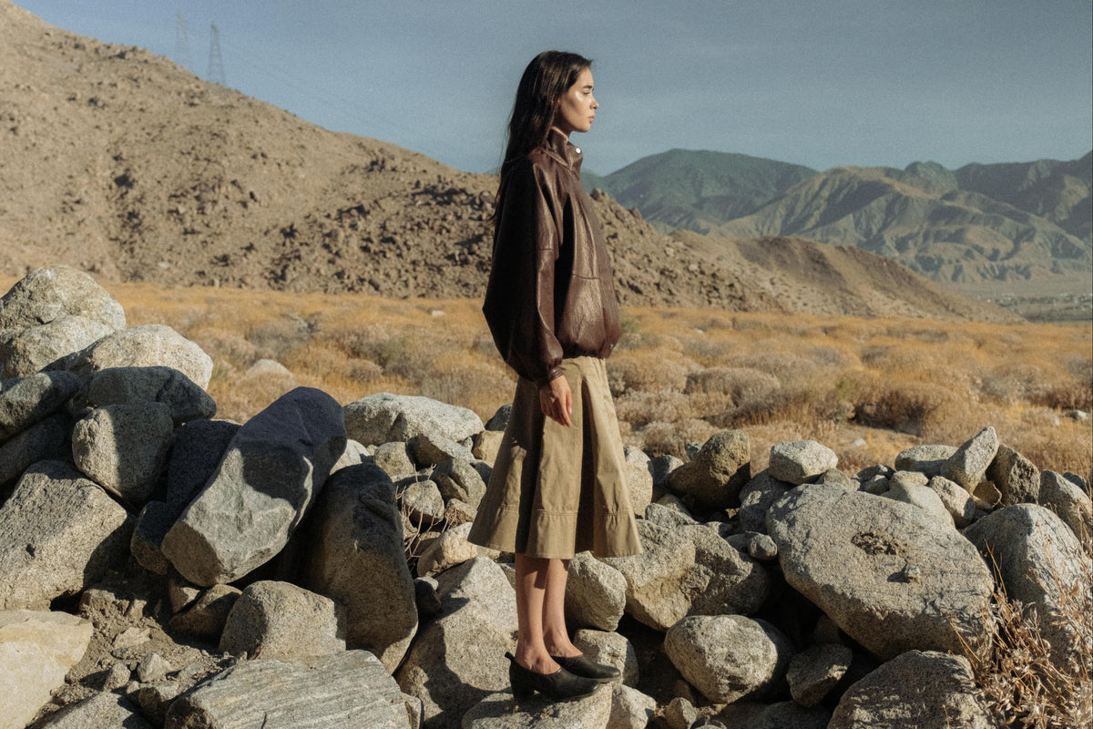 A woman stands on a pile of large rocks in a dry, mountainous desert landscape, wearing a dark brown leather jacket, a beige pleated skirt, and black flat shoes.